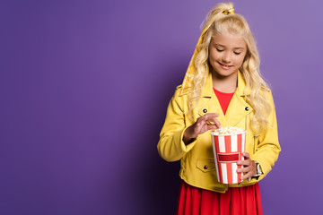 Fototapeta premium smiling kid holding bucket with popcorn on purple background