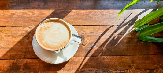 A hot cup of cappuccino coffee with milk foam on a wooden table under the light of the morning sun,...