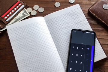 An open notebook on which the calculator lies and next to it are scattered dollars and rubles on a wooden brown background.