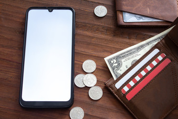 An oval smartphone with a white screen lies on a wooden table and next to it lies dollars and rubles with a leather wallet and a holder.