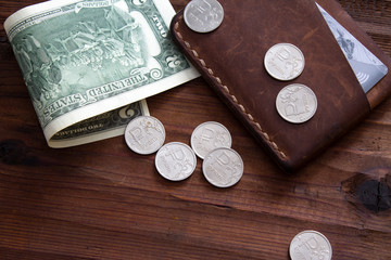 Dollar bill with scattered metal rubles on a wooden table next to a leather holder for money.