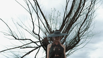 Portrait Of Pretty Blonde In Black Hat. Girl Stands Under Big Dry Tree.
