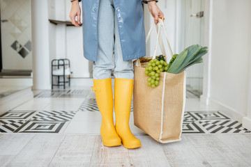 Fototapeta na wymiar Woman in yellow rubber boots and raincoat standing with shopping bag full of fresh food in the corridor. Woman coming home in rainy weather