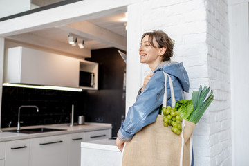 Young woman in blue coat coming home with shopping bag full of fresh vegetables and greens, walking on the kitchen