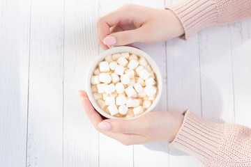 A Cup of hot chocolate and marshmallow in the hands of a young girl sitting at a light wooden table, top view .