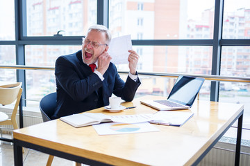 Adult male businessman working on a new project and looking at stock growth charts. Sits at a large window at the table. Looks at the laptop screen and drinks coffee.