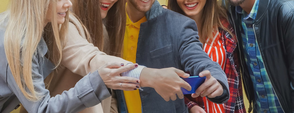 Group of friends standing by the wall background and enjoying each other- Teenagers using smartphone and smiling while trying to take a selfie- Technology concept