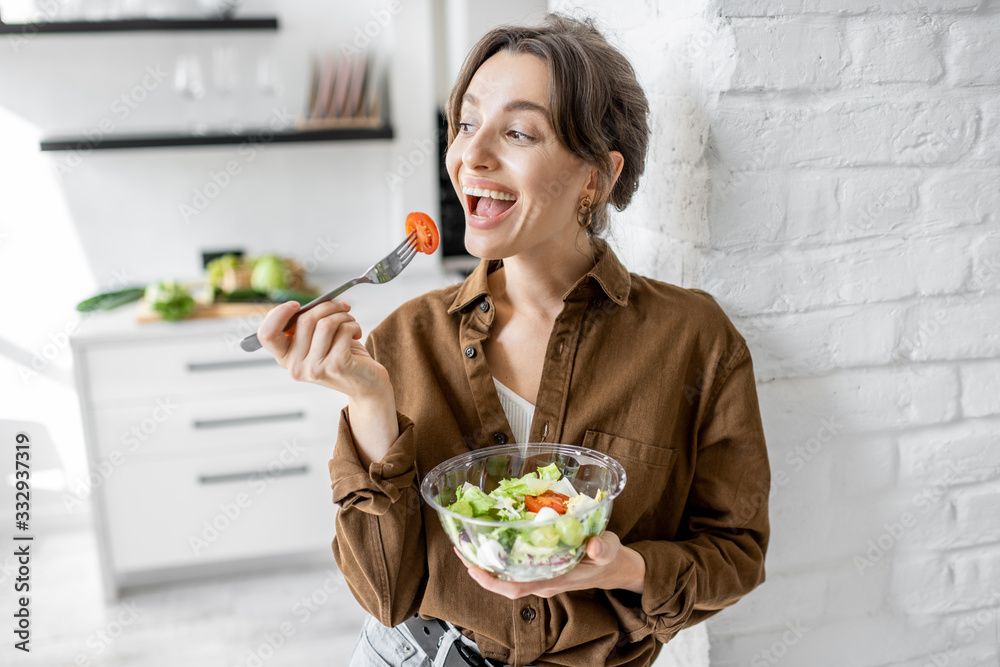 Wall mural portrait of a young and cheerful woman eating healthy salad on the kitchen at home. healthy eating, 