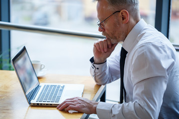 Male freelancer is working in a cafe on a new business project. Sits at a large window at the table. Looks at a laptop screen with a cup of coffee