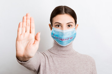 Portrait of a woman in medical mask with coronavirus text and showing stop gesture at white...
