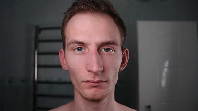 Playful handsome guy in the bathroom with his naked torso looks into the camera with a normal expression. View from the mirror. Close-up