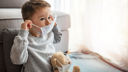 Child in home quarantine looking out of the window with his sick teddy bear wearing a medical mask...