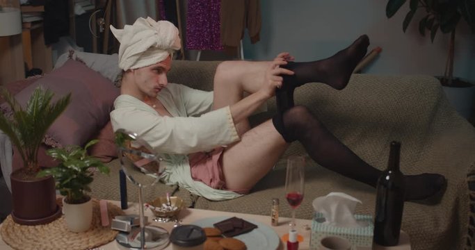 Drunky transgender man lying on sofa and putting on black tights. Relaxing young man transgender getting ready for party. Home background. Concept of people, beauty and leisure .
