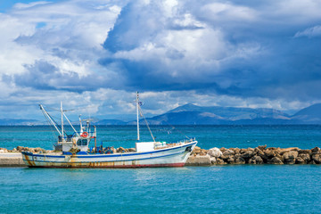 Beautiful landscape - sea bay with calm turquoise water, old fisher boat, stone breakwater, gray clouds on the sky and mountains on the horizon. 