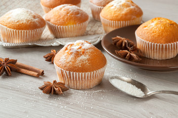 Freshly baked muffins are laid out on the kitchen table on a metal tray and in a plate. Cinnamon sticks and star anise and white sugar are scattered on the table. Sweet dessert.