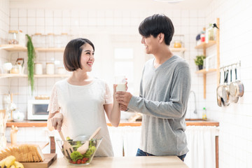 Asia new couple family stand in warm kitchen. Pregnant woman and husband hold glass of milk together. Girl cooks salad for man. They prepare healthy food in lovely home. Baby family pregnancy concept.