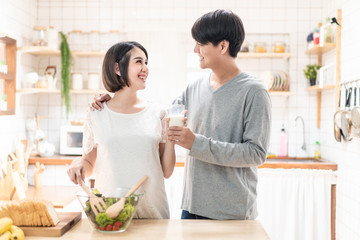 Asia new couple family stand in warm kitchen. Pregnant woman and husband hold glass of milk together. Girl cooks salad for man. They prepare healthy food in lovely home. Baby family pregnancy concept