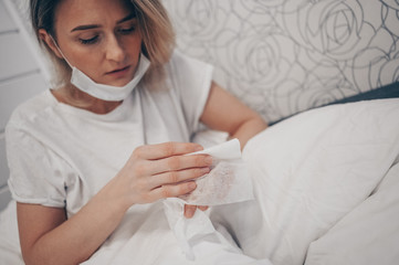 Woman in face protective mask lying in bed during coronavirus isolation home quarantine cleaning hands by hand sanitizer, using cotton wool with alcohol to wipe to avoid contaminating with COVID-19