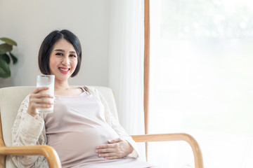 Young pretty Asian pregnant woman sits on armchair in front of white curtain holding milk and touching her tummy looks at camera with smile. Love care of pregnancy woman and baby health & food concept