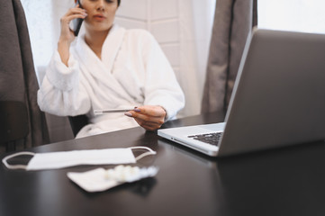 Fototapeta premium Sick woman speaking smartphone and working on a laptop during home quarantine isolation Covid-19 pandemic Corona virus. Distance work from home concept. Thermometer, pills and a medical mask on table
