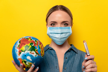 young woman in medical mask holding test tube with coronavirus blood sample and globe on yellow background