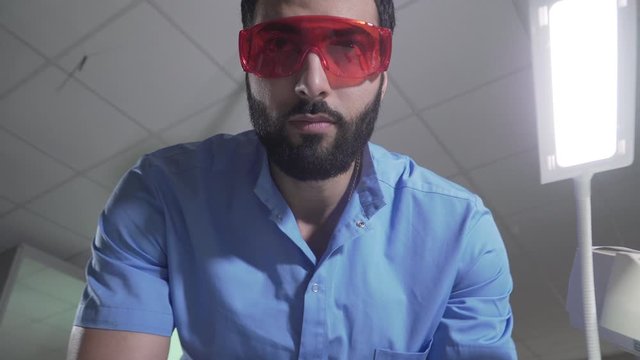 Serious Middle Eastern man in blue uniform and protective eyeglasses preparing for patient visit. Portrait of handsome bearded male dentist in office. Health care, medicine, occupation.