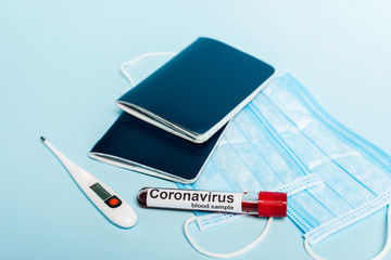 test tube with blood sample and coronavirus lettering near medical masks, thermometer and passports on blue background