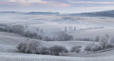 Panoramic Rural Landscape With Wavy Plowed Fields, Trees In Hoarfrost And Old Windmill In Morning Fog. Winter Arable Land. Beautiful Frosted Cultivated Field. Frozen Grass. Christams In Moravia, Czech