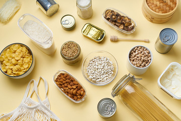 Creative flatlay with pantry staples. Glass jars with pasta, beans and chickpeas, canned goods, nuts and dried mushrooms in reusable containers. Top view pattern with basic products 