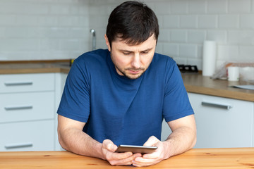Handsome bearded man in a blue T-shirt sits behind a wooden table and reads an electronic book against a blurred background of a modern kitchen