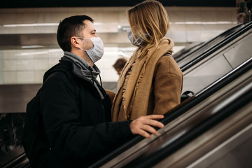 Loving man and woman looking at each other in medical masks on escalator in subway.