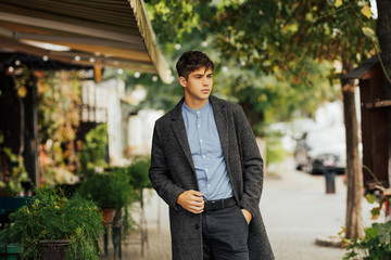 Handsome young serious man in elegant wear walking in the city while spending time outdoors. He dressed in autumn grey coat and blue shirt. Outdoor fashion male portrait.