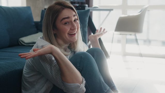 Close up view of funny young caucasian woman sitting on the floor near couch looking to the camera shrugs smiling enjoying having fun at modern room no stress concept slow motion