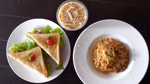 Top view of freshly cooked delicious spaghetti bolognese with parmesan cheese, toasted ham and cheese sandwiches and glass of coffee with ice, black wooden table. Concept of lunch time in restaurant.