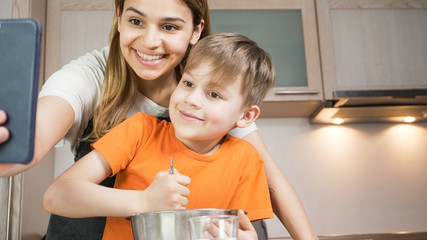 Beautiful mom and son cooking and making selfie at home. Happy family having a facetime video call. Unity, connection, home education concept.