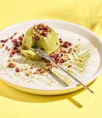 Matcha green tea, mint, lemon raw mousse cake on white plate and sunny yellow background. Contemporary minimalistic style. Raw Healthy Paleo green Avocado Cake. Tropical leaves and hard shadows around