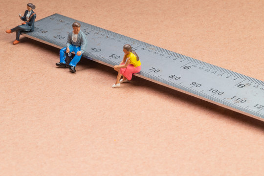 Miniature figures sit on a metal rule or ruler demonstrating the concept of social distancing to avoid transmission of the corona virus or other contagious illness