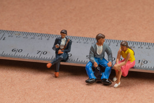 Miniature figures sit on a metal rule or ruler demonstrating the absence of social distancing leading to transmission of the corona virus or other contagious illness