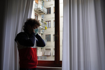 Europe, Italy, Milan - Man 40 years old at home in quarantine with mask looks out of the closed window to protect himself from the pandemic of n-cov19 Coronavirus                             