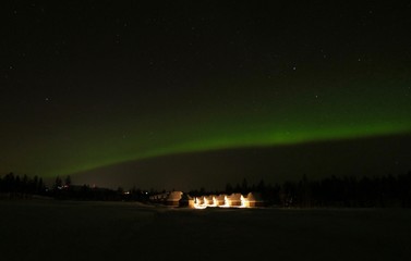 Naklejka na ściany i meble Green aurora borealis at night in snow landscape and cottages in background, Lapland, Finland