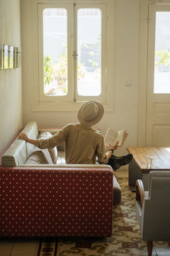 Young woman in hat reading book on sofa in room