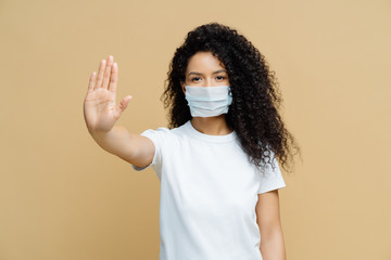 Serious dark skinned woman makes stop gesture, pulls palm towards camera, wears medical flu mask, asks stay at home not to spread coronavirus disease, prevents virus, dressed in white t shirt.