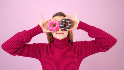 Adult female in read sweater smiling and keeping sweet round donuts near eyes looking at camera against pink background