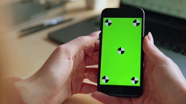 Close up view of european woman hands holding a green screen mobile phone or smartphone, tapping, scrolling, working on a desk with a laptop in the room. slow motion. Chroma key, tracking point