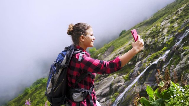 Pretty girl backpacker taking selfie in mountain. Woman in a red checkered shirt standing on the edge, recording video or sharing via smartphone while enjoying picturesque view waterfall from high