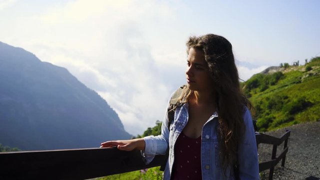 Attractive woman walking in mountain. Pretty girl in Jean jacket goes at the camera along tourist trail with wooden fence, enjoying picturesque view from top in sunny day