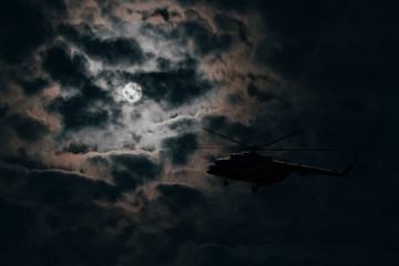 Silhouette of a military helicopter flying on a moonlit night against a cloudy dark sky
