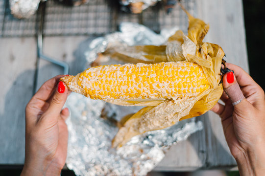 Young woman eating grilled Maize Cob in the open-air kitchen, against rustic wooden table