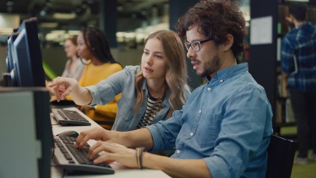University Library: Bright Caucasian Girl and Smart Boy Together Work on Computers, Chat, Discuss Class Assignment, Explain and Advice Each Other. Diverse Group of Students Exam Study Teamwork