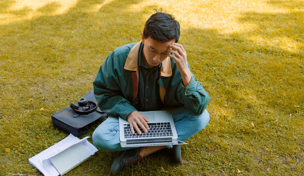 Thoughtful Asian student using laptop on lawn